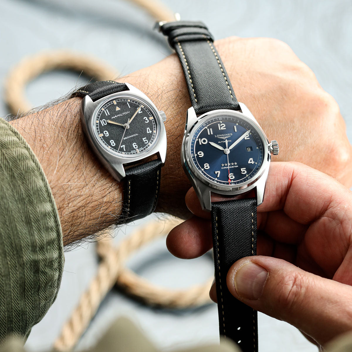 Hamilton watch and Longines watch fitted to black Mayday sailcloth style watch strap on a person's wrist against a blurred background