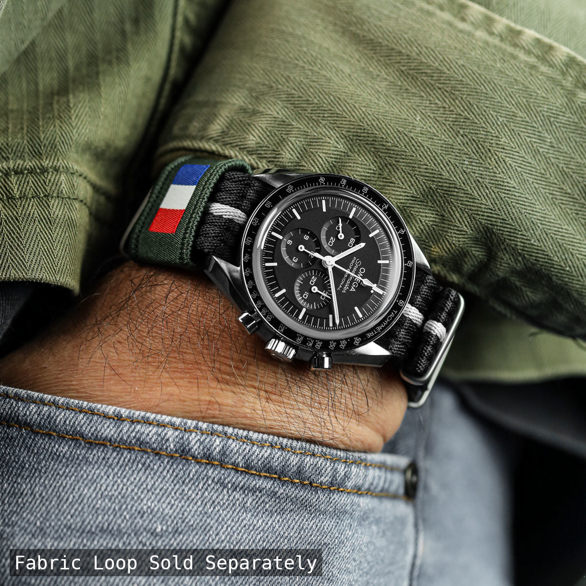 Omega speedmaster watch with a fabric strap featuring French flag design, worn on a wrist with green jacket and jeans.