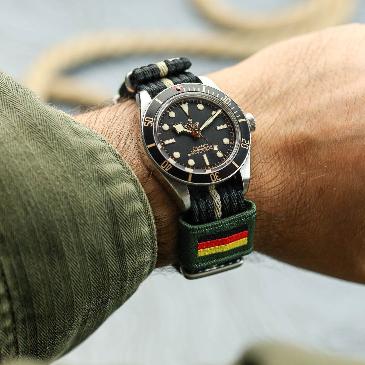 Tudor watch with a green strap featuring red and yellow stripes on a wrist, blurred background