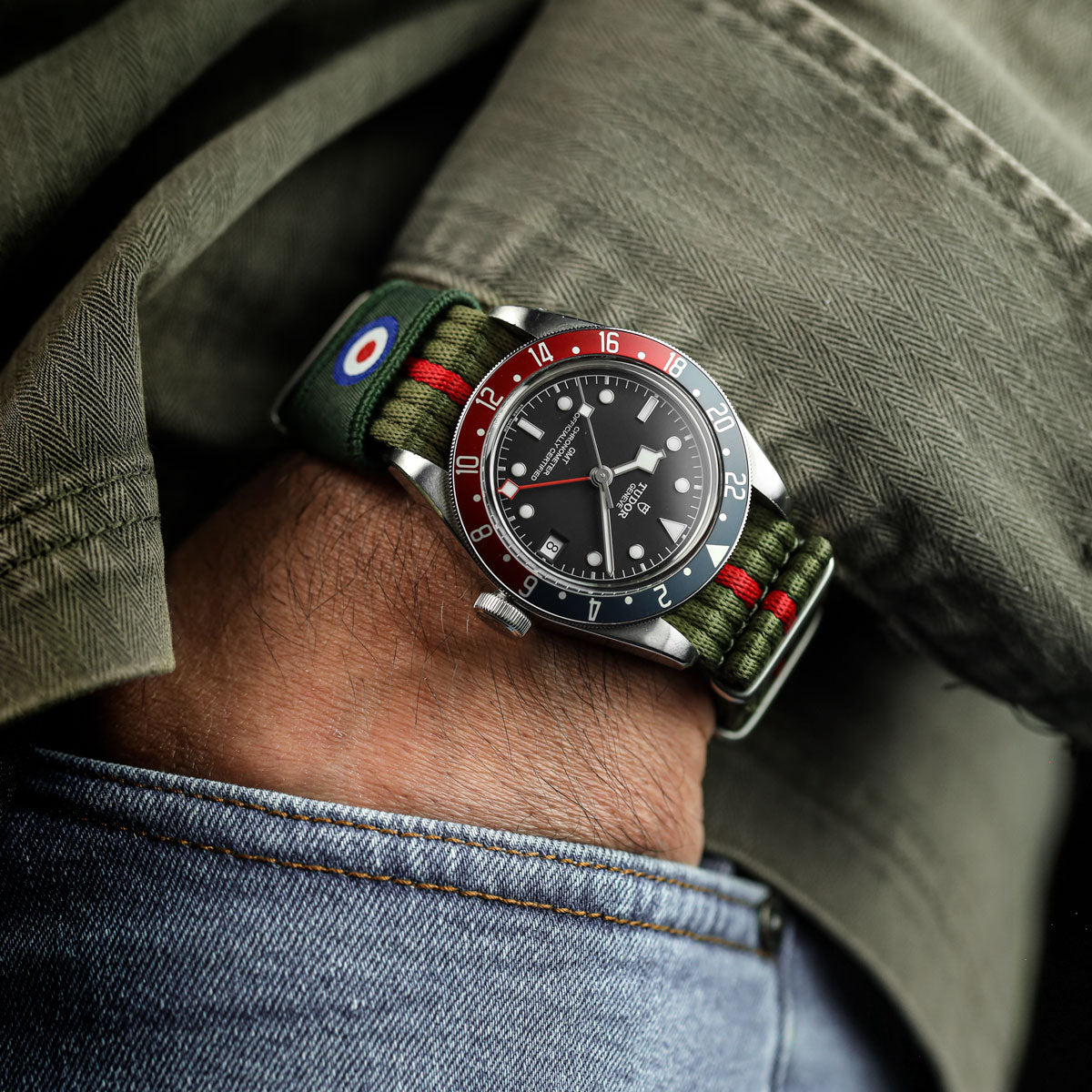Tudor watch with a green strap on a wrist, wearing a green shirt and blue jeans.