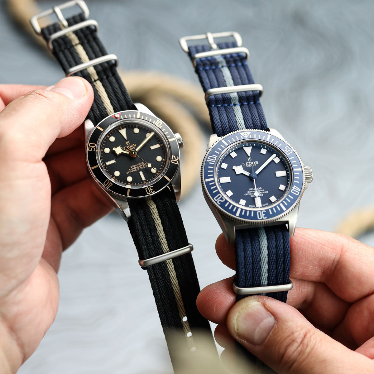 Two Tudor watches with fabric straps held by a person against a blurred background