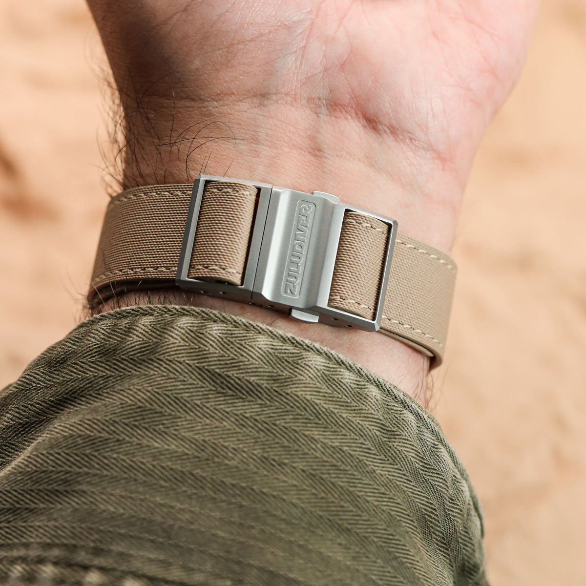 A close-up of a wrist wearing a beige woven strap watch with a silver-toned metal buckle engraved with the brand name 'ZULUDIVER'. The person is wearing an olive green herringbone shirt against a warm, sandy beige background.