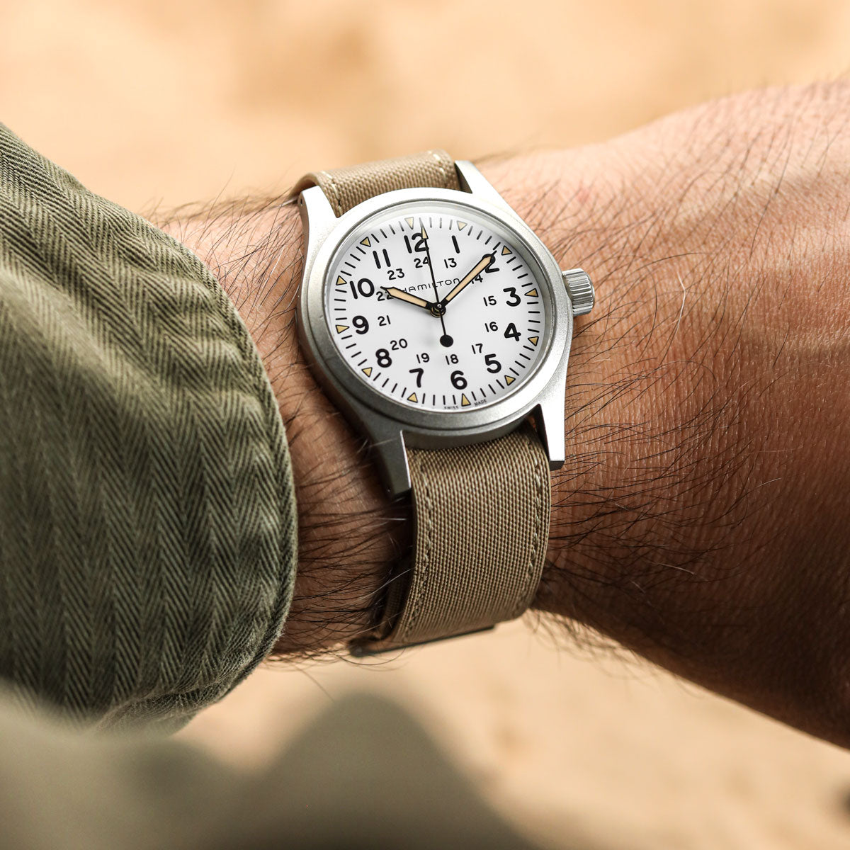 A wristwatch with a silver-toned metal case, white dial, and khaki canvas strap worn on a person's wrist.