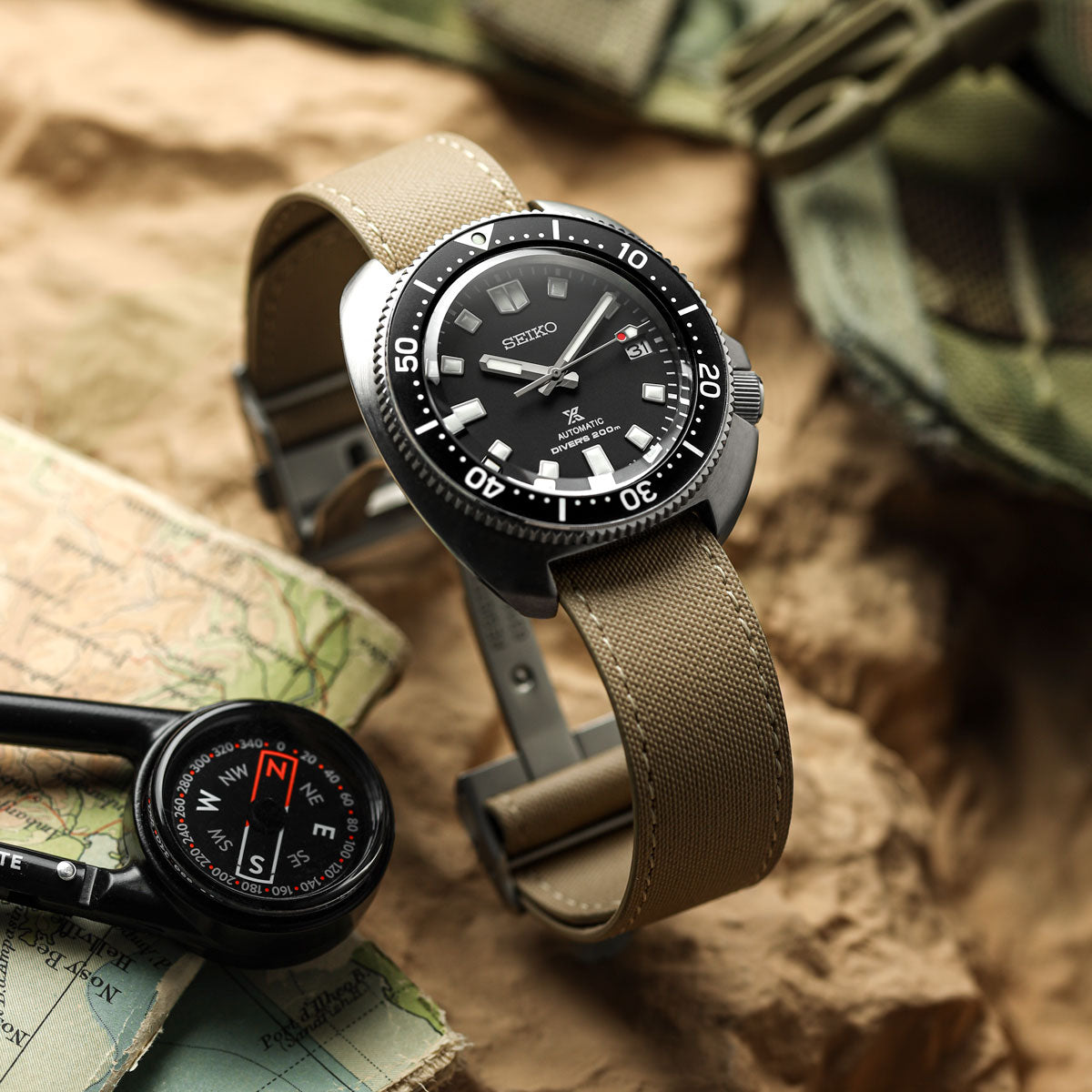 A Seiko dive watch with a black dial and a tan canvas strap, styled on rocks with a map, compass, and green fabric in the background.