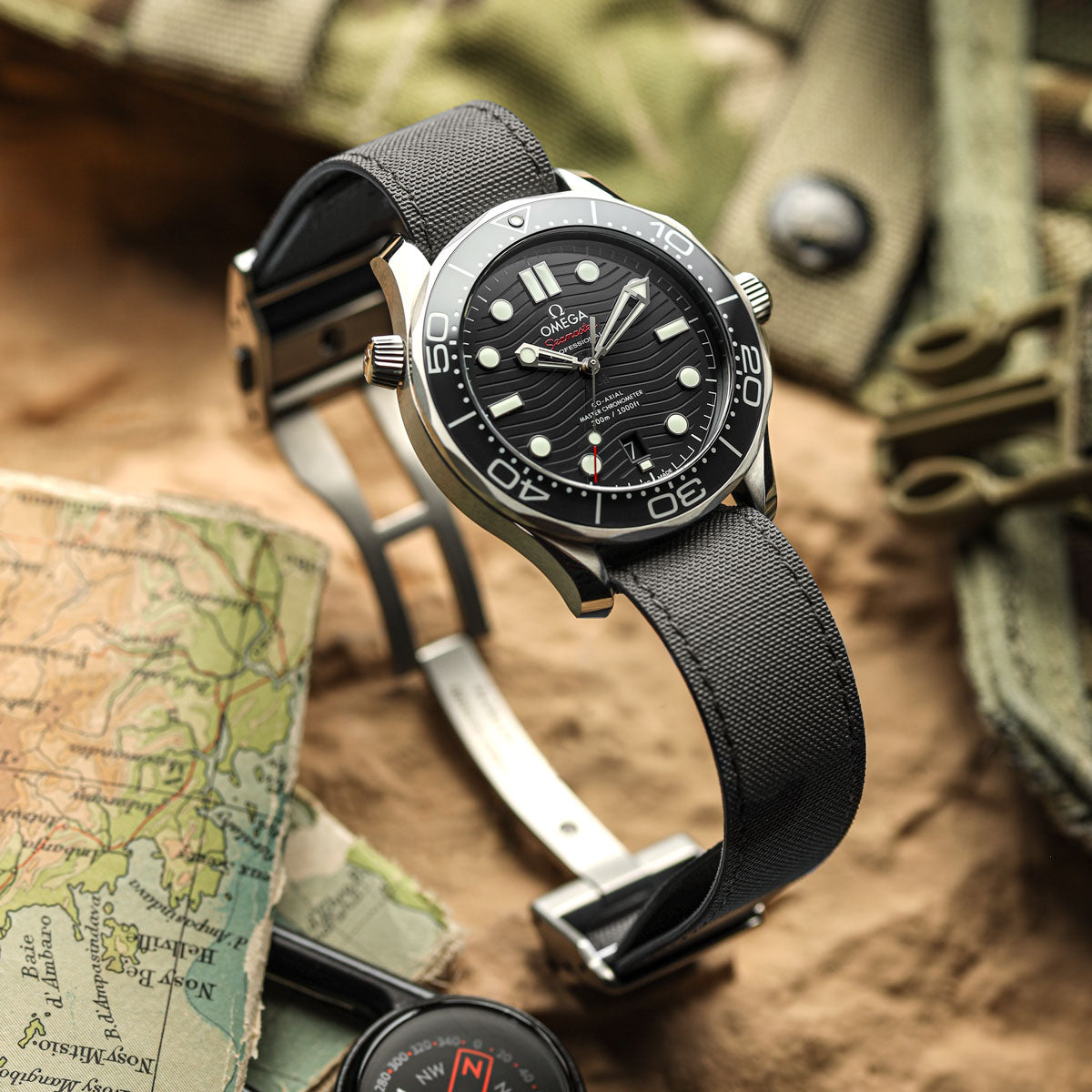 A wristwatch with a black dial, silver-tone stainless steel case, and black fabric strap, styled on a textured surface with a map, compass, and olive green gear in the background.