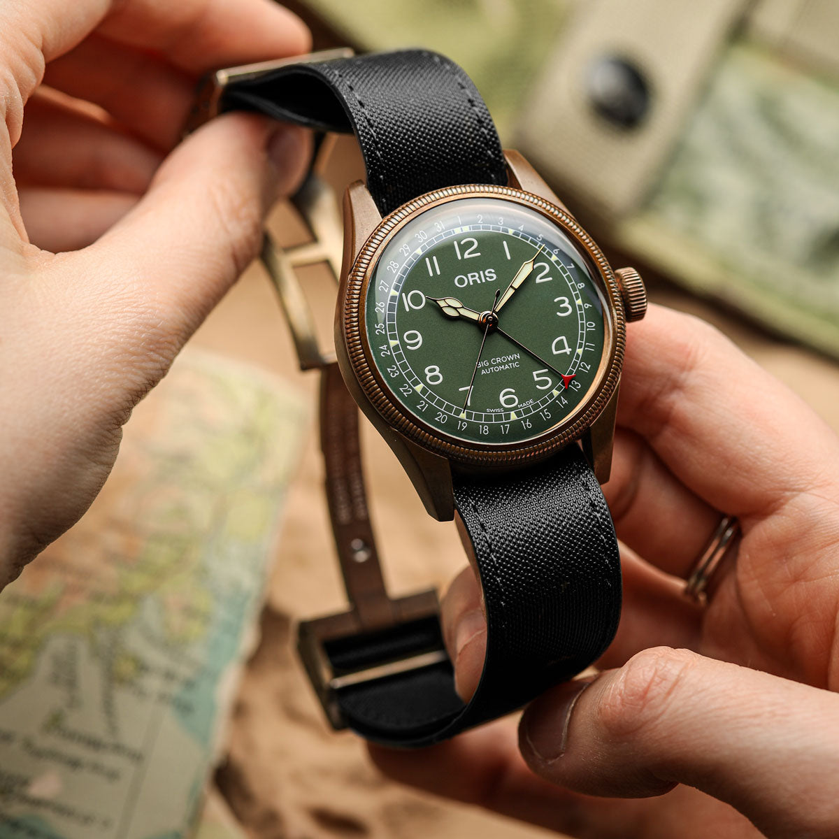 A close-up of a Oris wristwatch being held by two hands. The watch has a bronze-toned metal case, a dark green dial with white Arabic numerals and hands, and a black textured fabric strap with a black buckle.