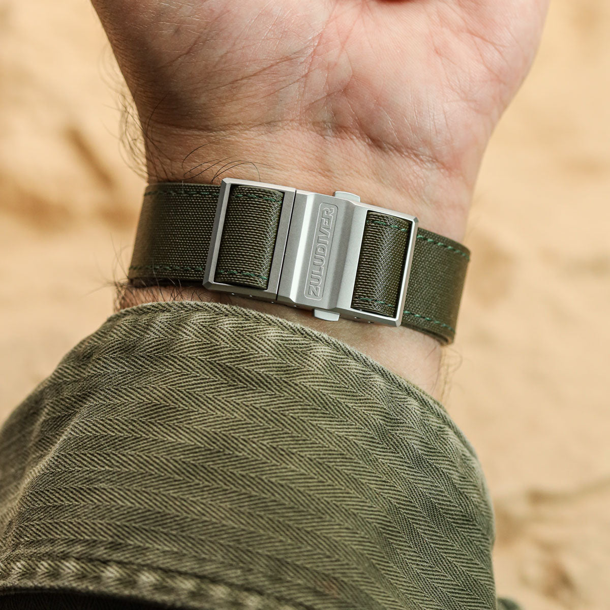 An olive green nylon sailcloth watch strap with a silver-toned metal buckle engraved with 'ZULUDIVER', worn on a person's wrist.