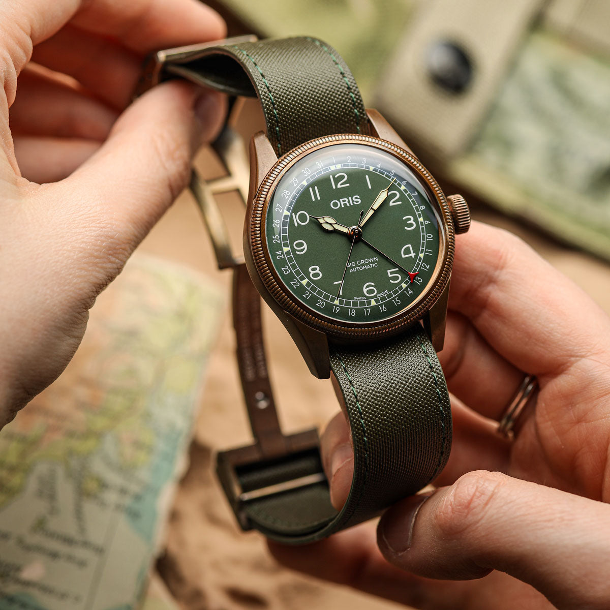 A close-up of an Oris Aviator watch with an olive green dial and matching green sailcloth fabric strap, held by two hands against a softly blurred background.