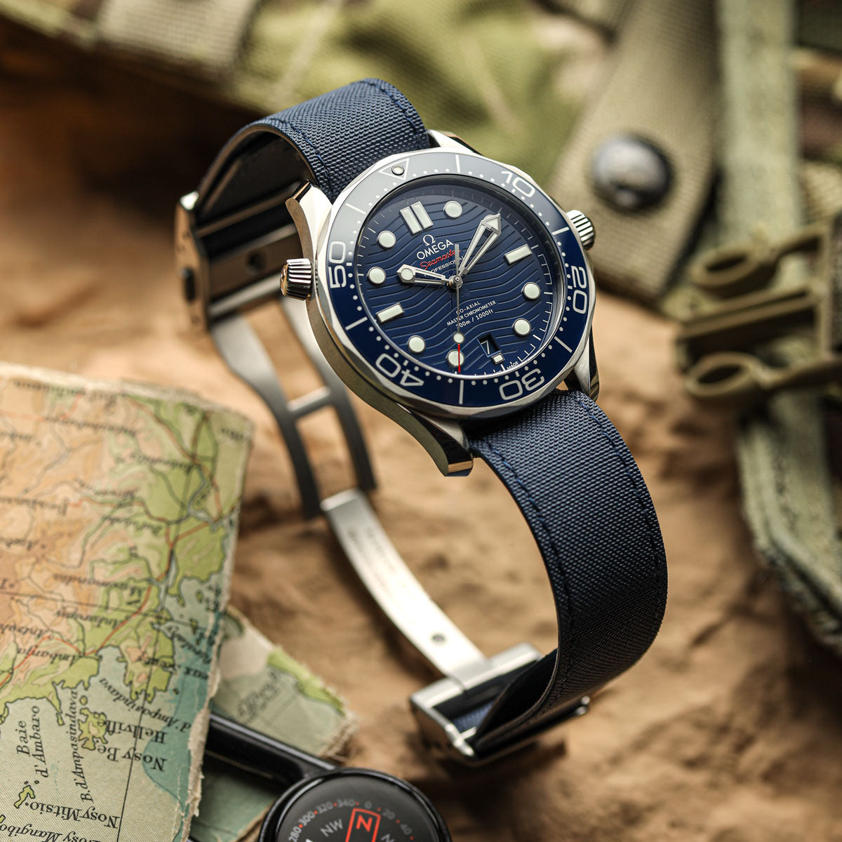 An Omega Seamaster wristwatch with a blue sunburst dial, silver-tone case, and dark blue Sailcloth fabric strap, styled on a rustic surface with a map, compass, and olive green gear in the background.