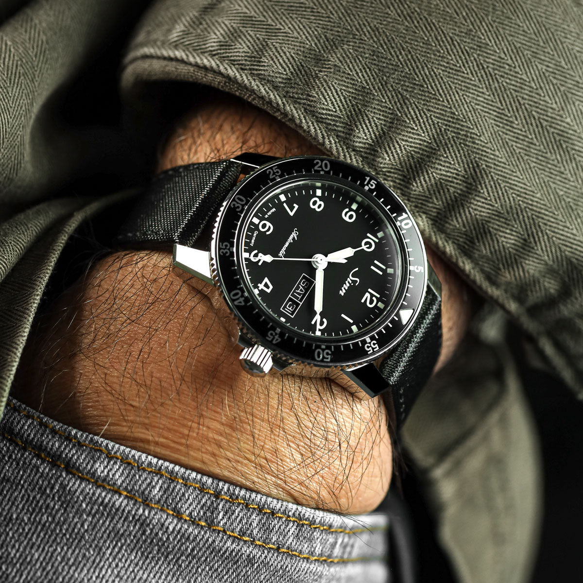 Sinn black wristwatch with a detailed face on a person&#39;s wrist, wearing a green jacket and gray jeans.