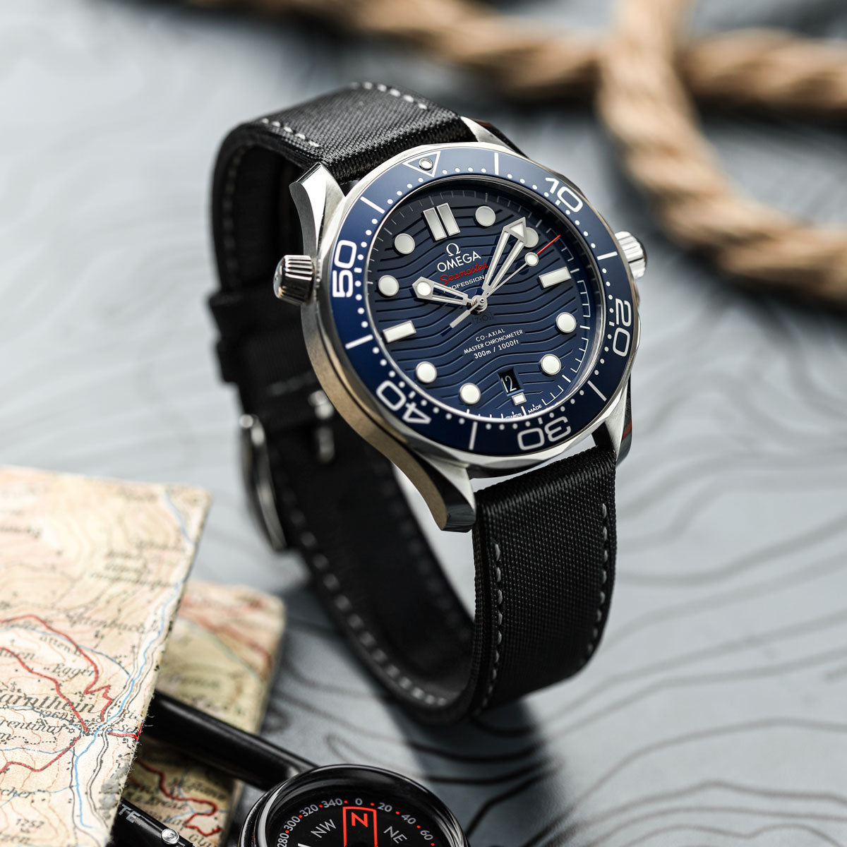 Omega seamaster watch with blue dial and black sail cloth watch strap with grey stitching on a textured surface with map and compass.