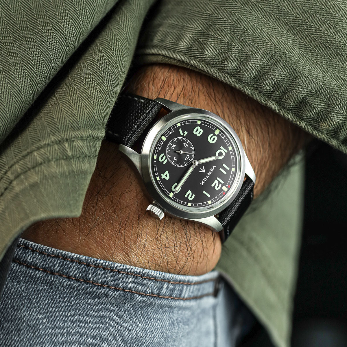 Close-up of a wrist wearing a Vertex black wristwatch with a detailed face, fitted to a Maverick sailcloth watch strap with grey stitching, against a green shirt and denim background.